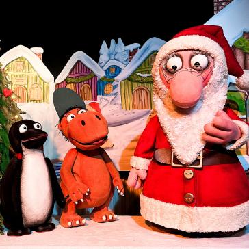 Weihnachtsszene: Santa Claus mit Renten, Pinguin, Igel und Nilpferd in Kostümen vor beleuchtetem Weihnachtsbaum und bunten Häusern.