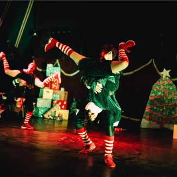 Tänzer in grünen und schwarzen Kostümen mit rot-weißen Strümpfen und Schuhen, dynamische Posen auf dunkler Bühne mit Weihnachtsbaum.