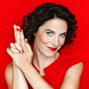 Person mit dunklen Locken und blauen Augen in rotem, ärmellosem Kleid, lehnt an roter Fläche und hebt eine Hand mit gespreizten Fingern, roter Hintergrund.