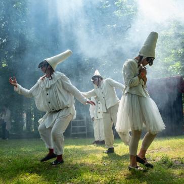Vier Figuren in weißen Pulcinella-Kostümen mit großen Hüten und Masken, drei tanzen auf Waldwiese im Sonnenlicht mit Nebel, eine im Hintergrund.