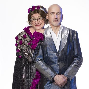 Zwei Personen vor hellem Hintergrund: links dunkles Kleid mit Silberpailletten und Blumenkopfschmuck, rechts glänzender silberblauer Anzug, Hände gefaltet.