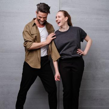 Ein Mann und eine Frau stehen vor einer grauen Wand. Beide lachen, der Mann trägt ein khakifarbenes Hemd, die Frau ein graues T-Shirt.