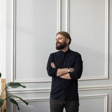Mann mit Mütze und Bart steht mit verschränkten Armen vor einer hellen Wand, neben einer Pflanze.