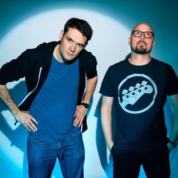 Auf dem Bild stehen zwei Männer, in einem Spotlight vor einem grünen Hintergrund. Der Mann rechts im Bild trägt eine Brille und hat eine Glatze. Er hand die Hände in den Hosentaschen und schaut gelangweilt nach oben. Zu seiner Rechten beugt sich der andere Mann nach vorne mit den Händen in den Hüften. Dabei schaut er uns herrausfordernd an.