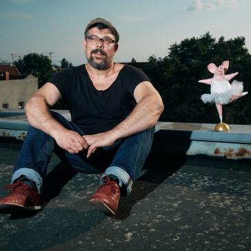 Ein Mann sitzt auf einem Dach und trägt ein schwarzes T-Shirt, Jeans und rote Schuhe. Neben ihm steht eine kleine, rosa Ballerina-Figur.