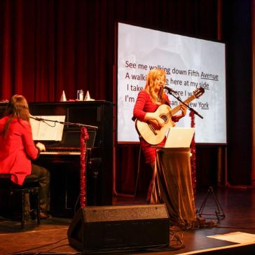 Zwei Musikerinnen führen auf einer Bühne auf. Eine sitzt am Klavier, während die andere Gitarre spielt und singt. Im Hintergrund ist ein großer Bildschirm mit projiziertem Songtext zu sehen. Die Bühne ist mit roten Vorhängen und festlichen Dekorationen geschmückt.