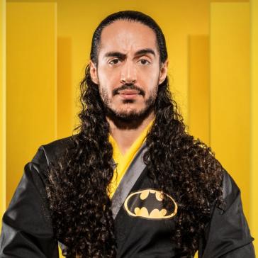 Ein Mann mit langen lockigen Haaren trägt ein schwarzes Oberteil mit Batman-Logo und steht vor einem gelben Hintergrund.