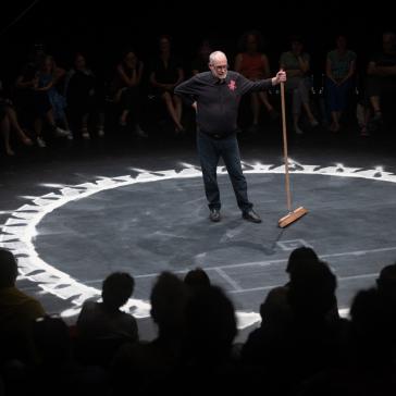  Ein älterer Mann steht in einem dunklen Theaterraum im Zentrum eines Kreises aus weißem Pulver und hält einen Besen, während das Publikum im Hintergrund sitzt.