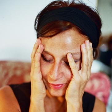 Eine Frau mit geschlossen Augen hält ihr Gesicht mit beiden Händen, sie trägt ein schwarzes Oberteil und ein schwarzes Haarband.