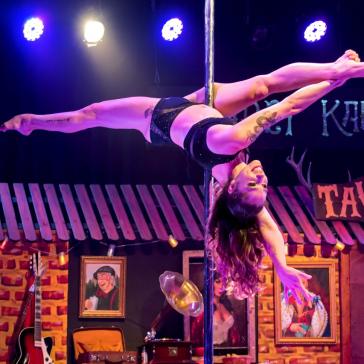 Eine Akrobatin, die in einem Spagat an einer Stange hängt, führt ihre Performance auf einer Bühne auf. Die Umgebung ist lebhaft gestaltet mit dem Schriftzug „Kabaret Kalashnikov“ und „Taverna“ im Hintergrund.