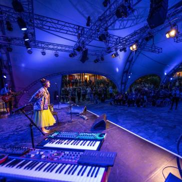 Eine Sängerin mit buntem Outfit steht barfuß auf einer Open-Air-Bühne unter einem Zelt, umgeben von Musikern. Ein großes Publikum sitzt im Halbrund, während blaue Lichter die Szene erhellen.
