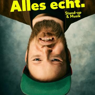  Das Bild zeigt eine Person, die auf dem Kopf steht. Es handelt sich um eine Nahaufnahme eines Mannes mit einem Bart und einer grünen Baseballkappe. Der Mann lächelt und schaut direkt in die Kamera. Der Hintergrund ist unscharf und in neutralen Grautönen gehalten.