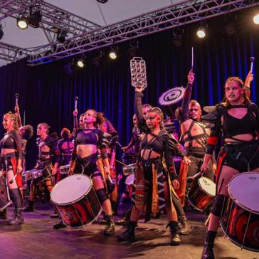 Das Foto zeigt eine Gruppe von Musikern auf einer Bühne. Die Musiker tragen markante, auffällige Kostüme in Schwarz und Rot. Einige von ihnen spielen große Trommeln, die sie vor ihrem Körper tragen, während andere verschiedene Perkussionsinstrumente halten. Sie alle wirken energetisch und engagiert, und einige heben ihre Instrumente oder Trommelstöcke in die Luft. Die Beleuchtung der Bühne ist dramatisch, mit bunten Lichtern, die die Szenerie hervorheben. 