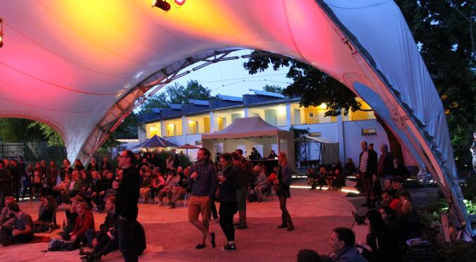 Eine abendliche Veranstaltung unter einem beleuchteten Zeltdach, viele Menschen sitzen und stehen, bunte Lichter, Gebäude im Hintergrund.