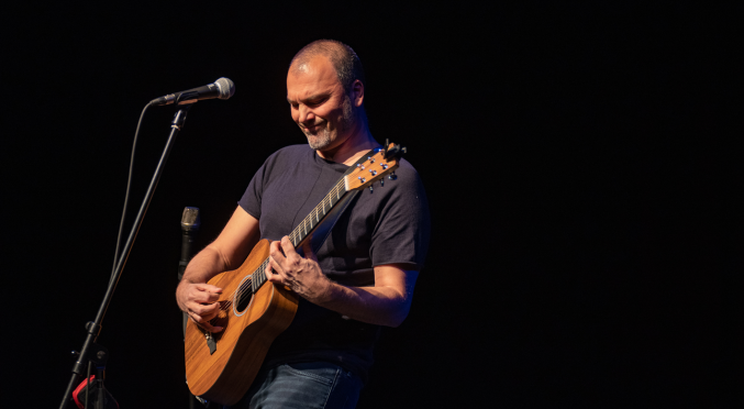 Ein weißer Mann mit sehr kurzen Haaren und einem kurzen, grauen Bart steht in leichter Rückenlage vor einem schwarzen Hintergrund. Er spiel eine Gitarre. Vor ihm ist ein Mikrofon auf einen Stativ. An der unteren Kante des Bildes ist noch ein Notenständer zu erkennen.