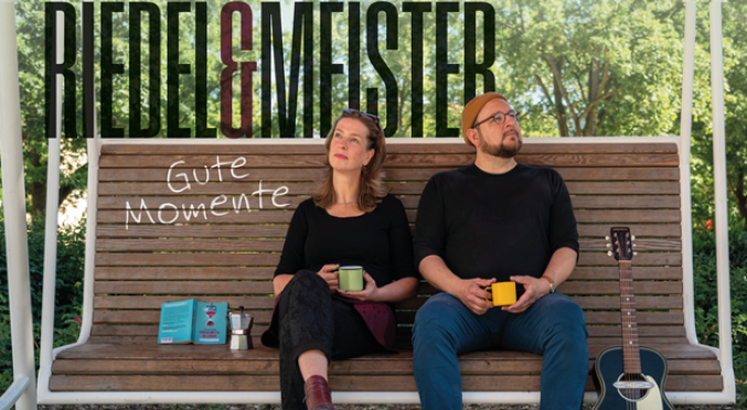 Ein Mann und eine Frau auf einer Schaukelbank, halten Tassen und schauen in die Ferne. Neben ihnen steht eine Gitarre und der Schriftzug "Gute Momente"