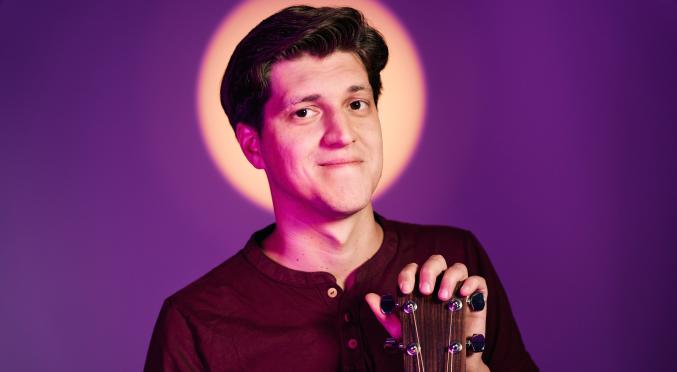  Das Bild zeigt einen jungen Mann, der eine Gitarre hält und sanft lächelt. Er ist in einem lila Licht, das ihn vor einem leuchtenden Kreis hervorhebt.