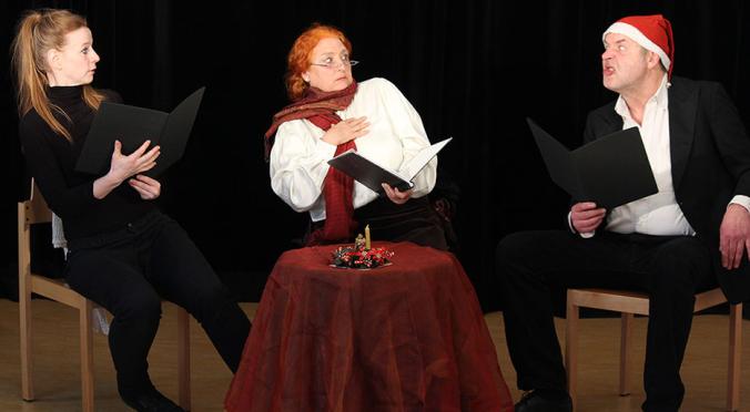  Das Bild zeigt drei Personen in einem Theaterstück. Sie sitzen um einen Tisch, halten schwarze Mappen in den Händen und zeigen lebhafte Gesichtsausdrücke.