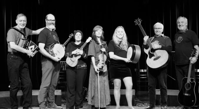 Das Bild zeigt sieben Musiker auf einer Bühne mit verschiedenen Instrumenten wie Banjo, Gitarre und Geige, alle in schwarzen T-Shirts, bereit für eine musikalische Darbietung