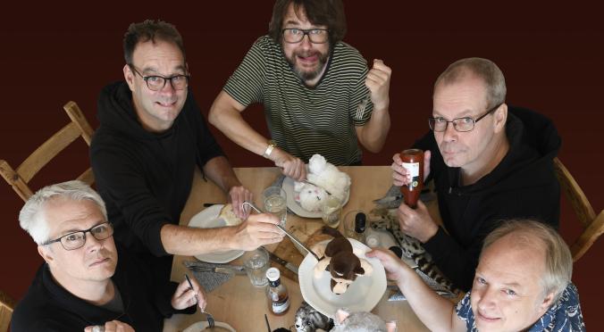  Das Bild zeigt fünf Männer an einem Tisch, die lächelnd in die Kamera blicken. Vor ihnen stehen Teller mit Stofftieren, und einer der Männer hält eine Flasche Ketchup.