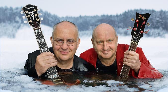 Das zeigt zwei Männer, die in eiskaltem Wasser stehen und Gitarren halten. Der Hintergrund zeigt schneebedeckte Bäume, was auf eine kalte, winterliche Umgebung hinweist.