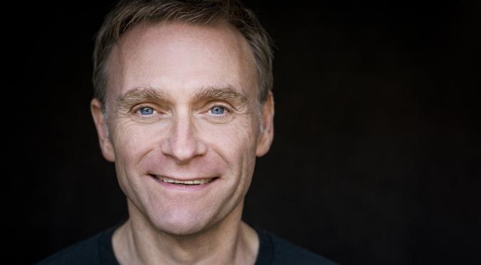  Das Bild zeigt einen lächelnden Mann mit blonden Haaren und blauen Augen vor einem dunklen Hintergrund. Er trägt ein dunkles Oberteil und wirkt freundlich und einladend.