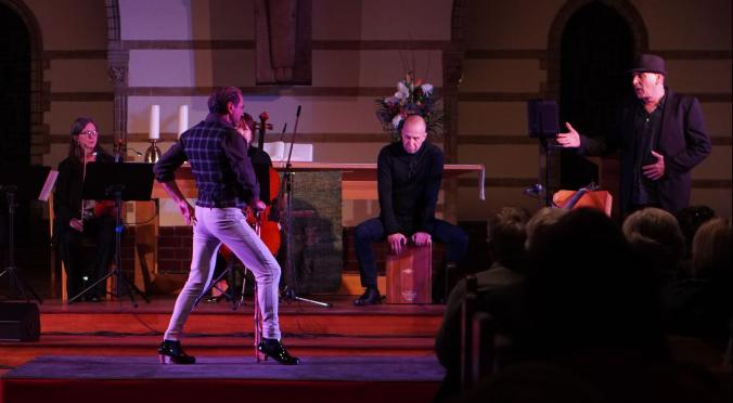 Ein atmosphärisches Bühnenbild in einer Kirche: Ein Mann mit Stock und hohen Absätzen steht im Rampenlicht, begleitet von Musikern und einem Sänger.
