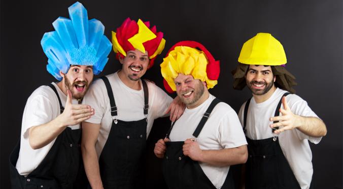 Das Bild zeigt vier Männer, die weiße T-Shirts und dunkle Latzhosen tragen. Sie haben bunte, auffällige Hüte, die an verschiedene Formen erinnern. Alle haben fröhliche Gesichtsausdrücke und scheinen Spaß zu haben. Wenn du mehr Details oder Informationen möchtest, lass es mich gerne wissen!