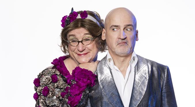  Eine humorvolle Szene: Zwei Personen posieren zusammen. Die eine Person trägt eine auffällige, glitzernde Robe mit violetten Blumen und eine Brille, lächelt liebevoll und legt den Kopf an die Schulter der zweiten Person. Die zweite Person, mit kahlköpfigem Kopf, trägt einen glänzenden silbernen Anzug und schaut skeptisch zur Seite.