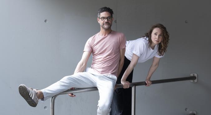 Ein Duo in legerer Kleidung posiert spielerisch auf einem Geländer. Eine lockere und kreative Atmosphäre vor einer minimalistischen Betonwand.