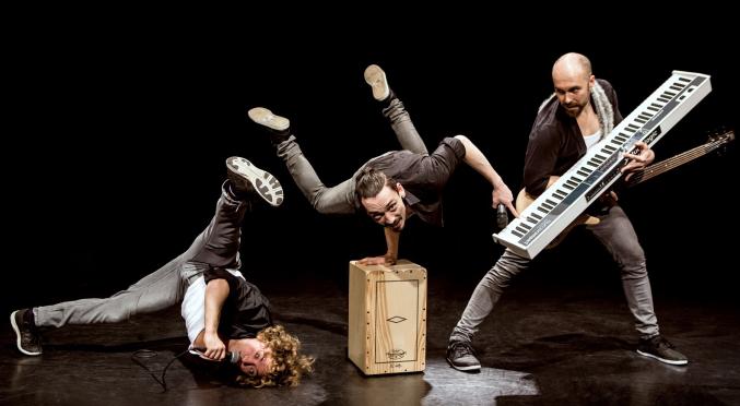 Dynamische Szene einer Performance-Gruppe: ein Akrobat balanciert einhändig, ein Musiker hält ein Keyboard und ein Sänger liegt am Boden, Mikrofon in der Hand.
