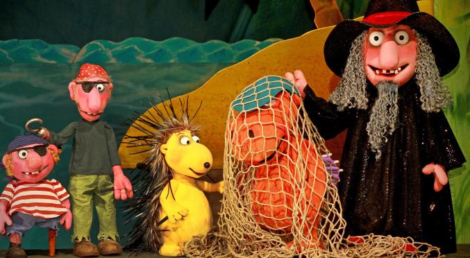 Eine Puppentheater-Szene mit Drachen, Zauberer, Piraten und einem gelben Stacheltier. Farbenfrohes Bühnenbild mit Meer und Insel im Hintergrund.
