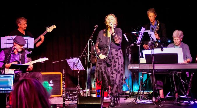 Das Bild zeigt eine Band, die auf einer Bühne spielt. Es sind mehrere Musiker zu sehen: eine Sängerin im Mittelpunkt, ein Gitarrist, ein Bassist, ein Saxophonist und eine Keyboardspielerin.