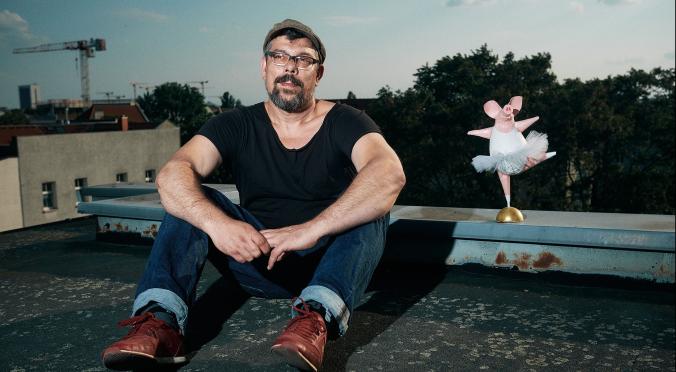 Ein Mann sitzt auf einem Dach und trägt ein schwarzes T-Shirt, Jeans und rote Schuhe. Neben ihm steht eine kleine, rosa Ballerina-Figur.