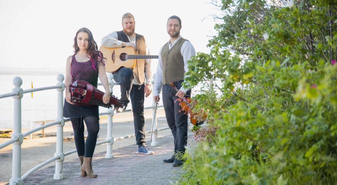 in Trio steht auf einem Gehweg am Meer. Links eine Frau mit einer Drehleier, rechts zwei Männer, einer mit Gitarre und der andere mit Geige.