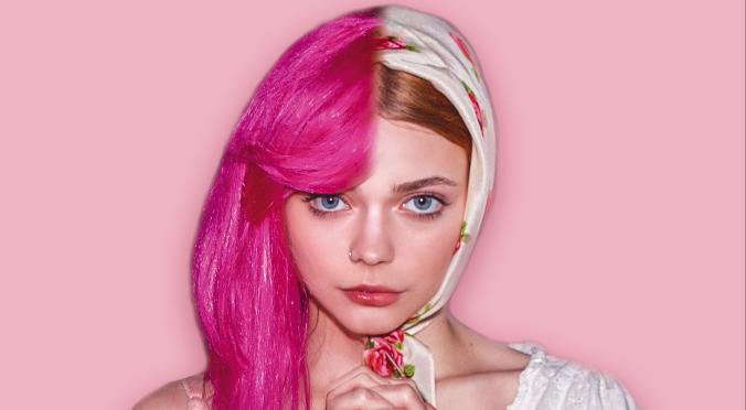 Das Bild zeigt Mädchen, dass links pinke Haare und rechts ein buntes Kopftuch trägt. Sie hat einen ernsten Gesichtsausdruck und hält ihre Hände vor dem Körper zusammen. Der Hintergrund ist in sanften Rosa-Tönen.