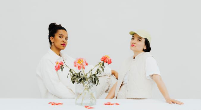 Das Bild zeigt zwei Frauen mit unterschiedlichen Stilen, die entspannt vor einem hellen, neutralen Hintergrund sitzen. Zwischen ihnen steht eine Vase mit Blumen. Beide blicken selbstbewusst in die Kamera.