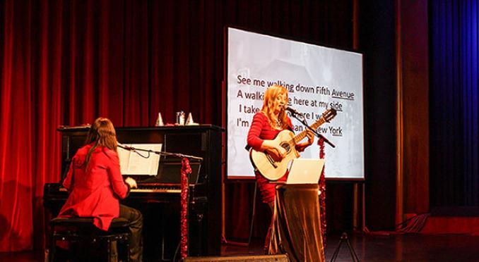 Zwei Musikerinnen führen auf einer Bühne auf. Eine sitzt am Klavier, während die andere Gitarre spielt und singt. Im Hintergrund ist ein großer Bildschirm mit projiziertem Songtext zu sehen. Die Bühne ist mit roten Vorhängen und festlichen Dekorationen geschmückt.