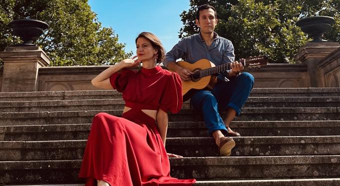 Eine Frau in einem roten Kleid und ein Mann mit einer Gitarre sitzen auf einer Steintreppe im Freien. Der Hintergrund zeigt sonnige Bäume und Architektur.