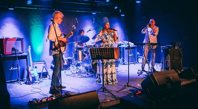 Eine Band spielt auf einer Bühne in einem Club. Links ein Gitarrist, in der Mitte eine Sängerin, rechts ein Trompeter, im Hintergrund ein Schlagzeuger.