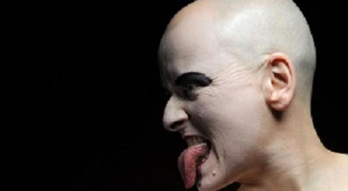 Das Bild zeigt eine Person mit einer glatzköpfigen Frisur und auffälligem Make-up. Sie hat einen provokanten Gesichtsausdruck und streckt die Zunge heraus, während sie mit einer Hand eine ausdrucksstarke Geste macht. Ein rotes Kreuz auf der Brust deutet möglicherweise auf eine künstlerische oder symbolische Bedeutung hin. Der Hintergrund ist schwarz, was die Figur und die Gesichtszüge hervorhebt.
