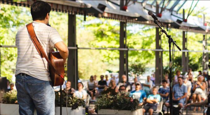 Das Bild zeigt einen Musiker, der mit einer Gitarre auf einer Bühne steht und ein Publikum ansieht. Der Hintergrund ist grün und hell, mit Bäumen und Pflanzen, was eine lebendige Atmosphäre schafft. Das Publikum ist in einer entspannten Position und scheint der Darbietung aufmerksam zuzuhören.
