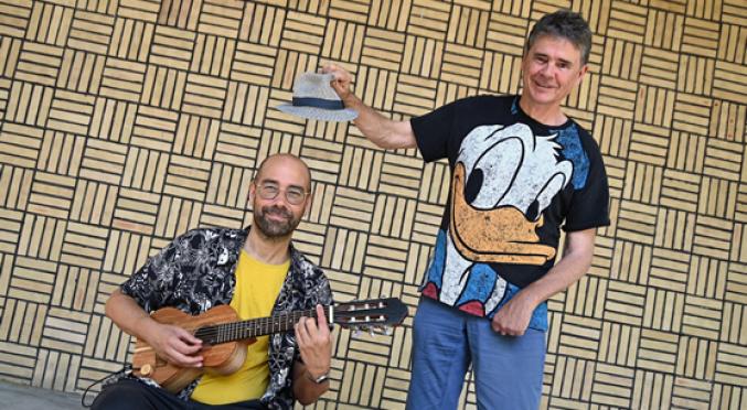 Ein Mann mit Brille spielt Gitarre, während ein anderer daneben steht, eine Mütze hebt und ein T-Shirt mit Donald-Duck-Motiv trägt. Hintergrund mit Muster.