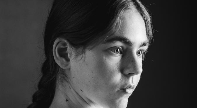 Das Bild zeigt ein Schwarz-Weiß-Porträt einer Person mit langen, geflochtenen Haaren. Die Person schaut mit intensivem Blick zur Seite, und das Licht betont die Gesichtszüge stark durch einen hohen Kontrast zwischen Licht und Schatten. Der Hintergrund ist dunkel und schlicht, wurde die Aufmerksamkeit auf das Gesicht und den Ausdruck der Person gelenkt. Die Aufnahme hat eine künstlerische und dramatische Wirkung.