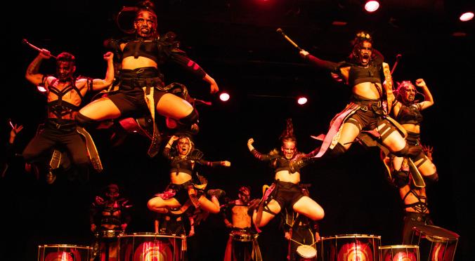 Trommelgruppe in Kriegerkostümen springt dynamisch in die Luft. Rote Bühnenbeleuchtung betont die Energie der Performance. Im Hintergrund stehen weitere Trommler mit bemalten Trommeln.
