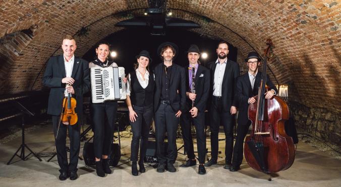 Eine Band aus sieben Menschen in eleganter Kleidung steht in einem Backsteingewölbe. Sie halten Instrumente wie Geige, Akkordeon, Klarinette und Kontrabass und lächeln in die Kamera.