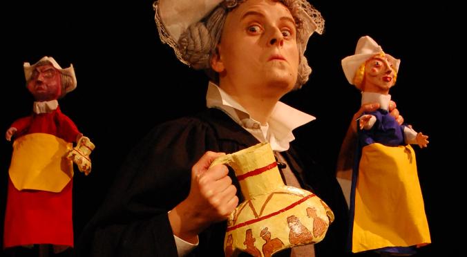 Eine Person in Kostüm mit Haube hält zwei Puppen und einen zerbrochenen Krug. Dramatische, historische Szene auf dunklem Hintergrund.