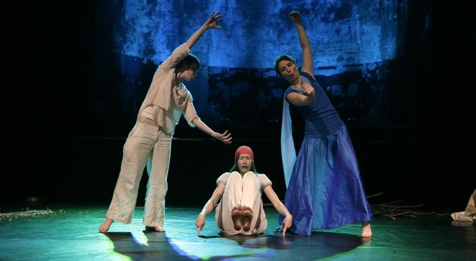 Das Bild zeigt drei Tänzer auf einer Bühne, die eine ausdrucksstarke Choreografie aufführen. Zwei der Tänzer sind stehend und bewegen sich anmutig, während der dritte auf dem Boden sitzt. Die Kostüme sind in Weiß- und Blautönen gehalten. Der Hintergrund ist dunkel, mit blauen Akzenten, die eine künstlerische und eindrucksvolle Atmosphäre schaffen.