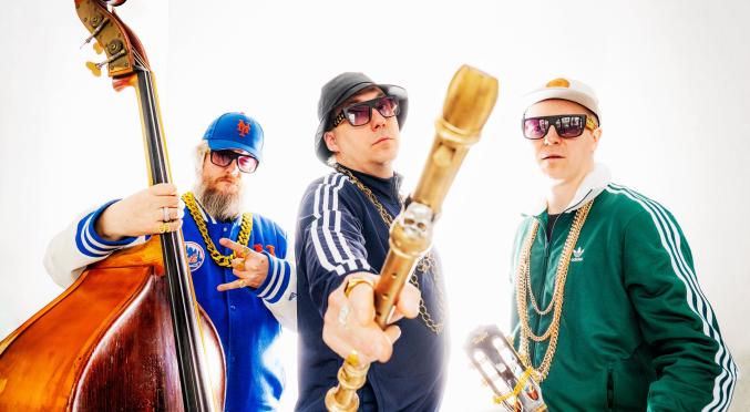 Drei Männer in Sportkleidung und Goldketten posieren cool mit Musikinstrumenten, darunter ein Kontrabass und eine Gitarre. Einer hält eine große Blockflöte in die Kamera.