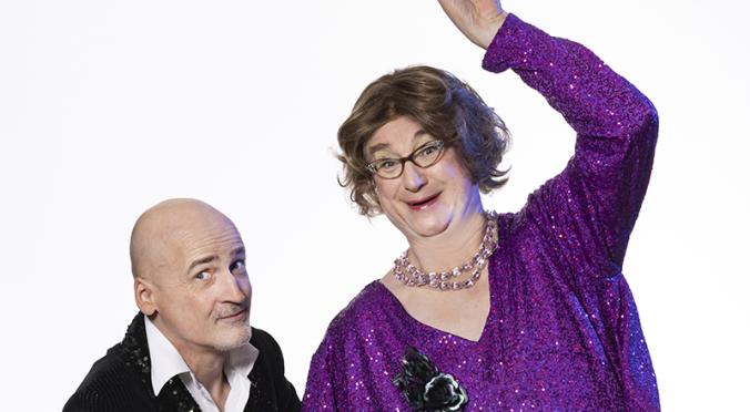 Zwei Personen posieren fröhlich: links ein Mann in schwarzem Glitzeranzug, rechts eine Person in lila Glitzerkleid mit Perlenkette, Brille und erhobener Hand in verspielter Pose.